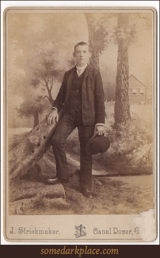 A young man standing next to a prop of a fallen log. He is wearing a dark suit and has close cropped hair and ears that stick straight out. He is holding a hat in his left hand.