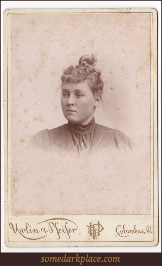 A curly haired young woman with her hair pulled up into a topknot. It appears there is a bow in her hair or some other ornament. She has round cheeks and her ears are pierced.