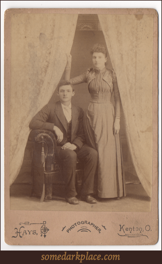 A young couple. The man is seated, the woman stands at his right to the left. Her hand is holding onto a curtain. They are both wearing formal wear. This photo is a studio shot.