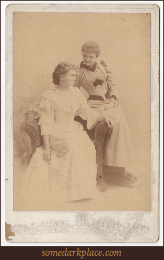 Two attractive young women. One fully seated and the other leaning into the armrest of this chair. One woman is looking at the other affectionately. Both women are wearing formal dresses.