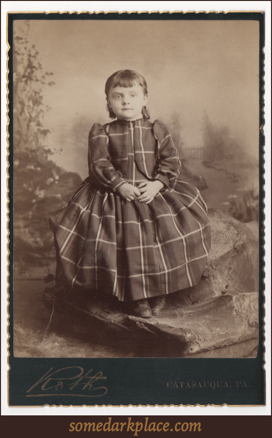 A young girl in a full sleeved full length dress. She has sausage curls and shoulder length hair. Her bangs are straight and evenly trimmed. She is seated on a prop rock in a studio shot in front of a painted background.