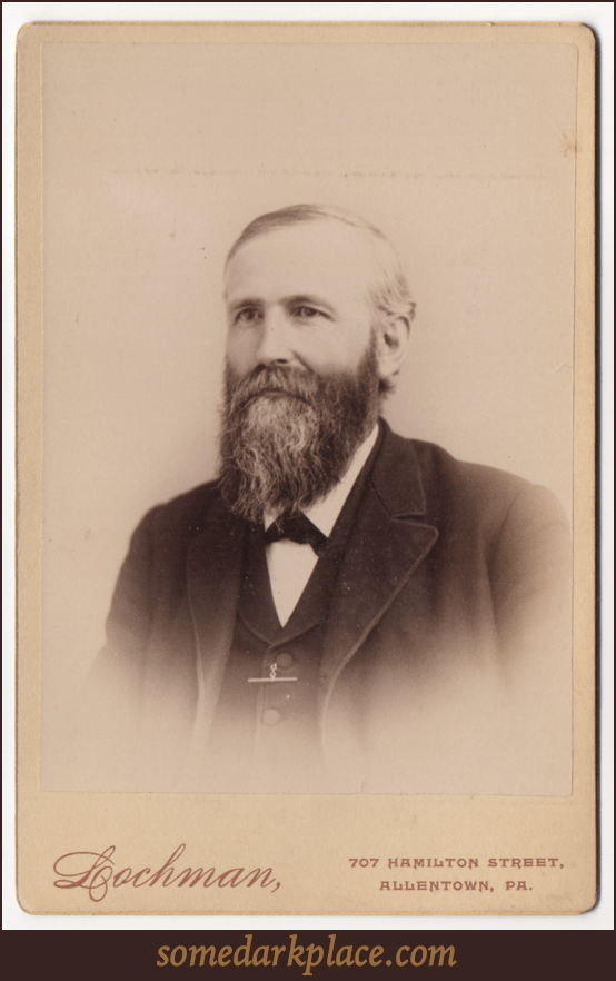 An older bearded gentleman wearing a suit coat and undershirt. He is wearing a dark tie of some sort. He has a watch chain or some other functional appealing jewelry. His beard and hair are starting to gray.
