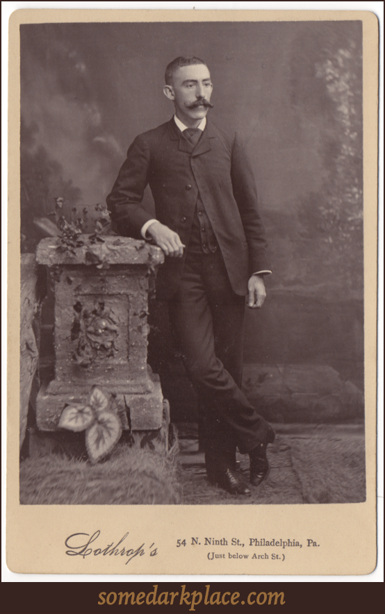 A man in a full dark suit leaning against a prop of a garden wall. He has on a light colored dress shirt and dark shoes with a shine to them. His mustache is large and impressive and well groomed.