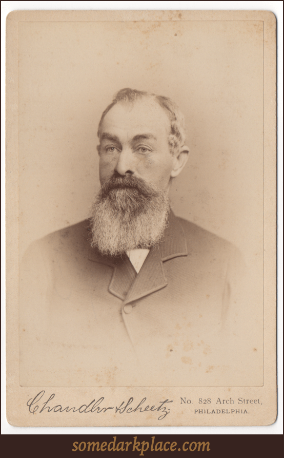 An older bearded gentleman wearing a suit coat and undershirt. He does not appear to be wearing a tie, but this could be obscured by his beard.