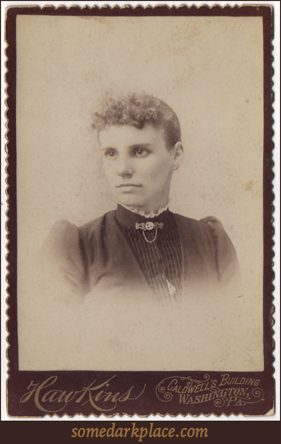 A curly haired young woman with a high collared dress with lace trim and a clasp and chain at her throat. Her dress is pleated in the front and has a piece of jewelry hanging in the front.