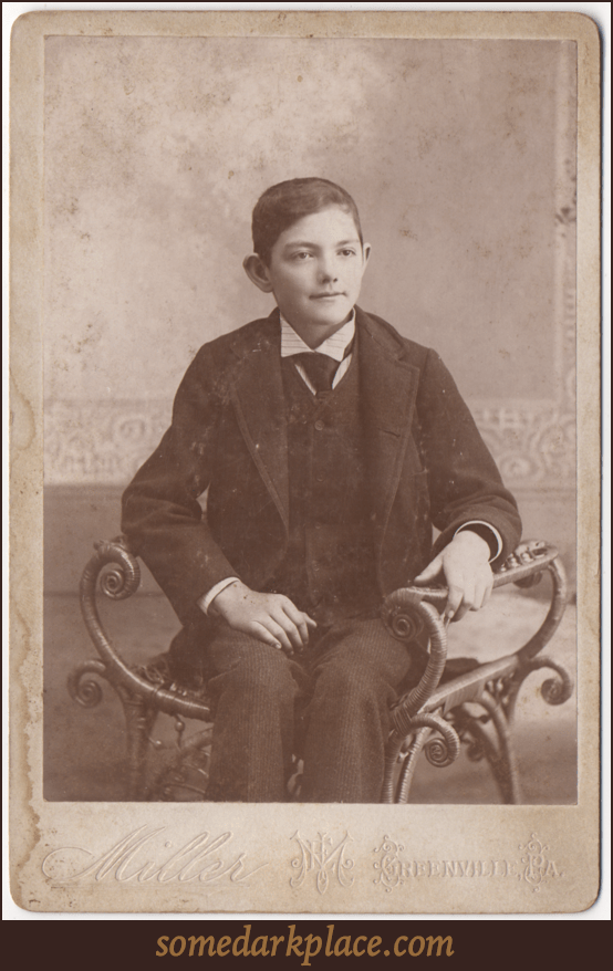 A young boy seated in an elaborate chair or bench without a back. He is dressed in formal attire, wearing a suit coat and tie. His hair is short, trimmed, and neatly combed. His ears stick out.