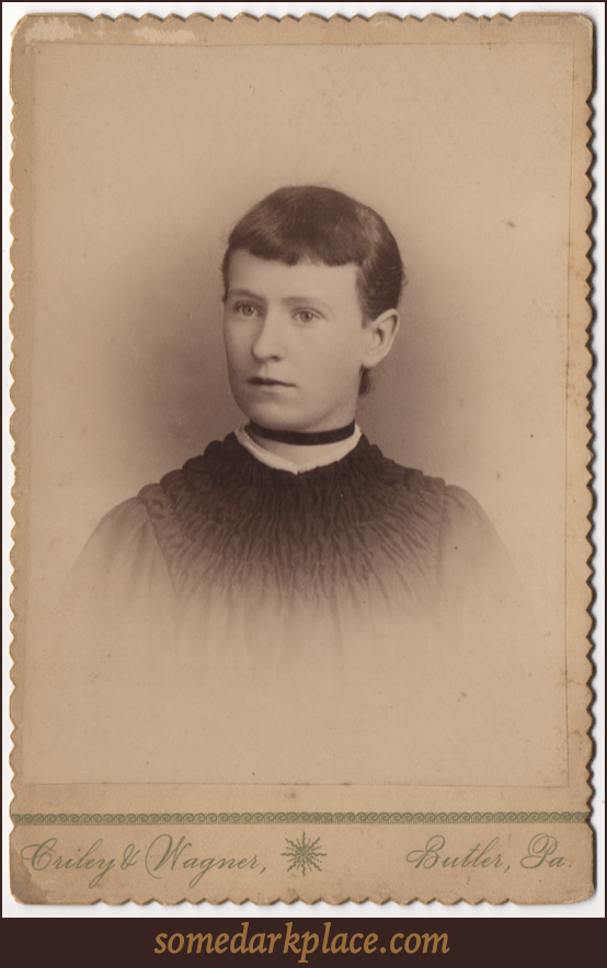 A short haired young woman with dark straight hair parted on the left. She is wearing a dress and does not have pierced ears. She wears a ribbon around her throat like a choker.