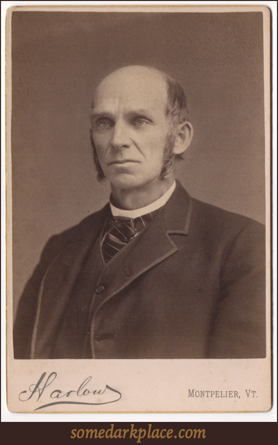 A bald older gentleman with log sideburns. He is wearing a suit coat with a matching undervest. He wears a tie and has a round collar that could possibly denote clergy.