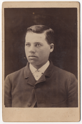 A handsome young man. He's wearing a suit coat and a white collared shirt. He appears to be wearing a tie of some sort. His hair is parted on the left.