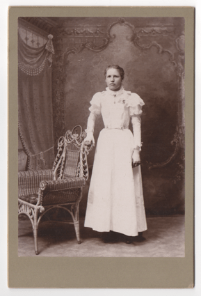 A young woman looking displeased and like she does not want her picture taken at all. She is wearing a full white gown with puffy shoulders. She also has fingerless gloves with piping on the back of the hands.