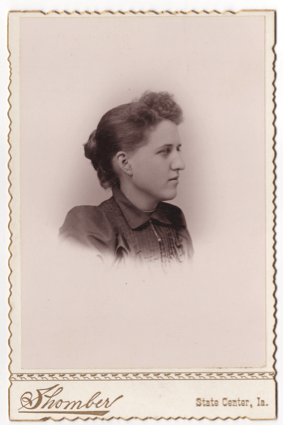 A bust image of a young woman in a dark dress or blouse with a small dangling pendant. Her hair is done in a bun at the back of the head and is loose curls in the front.