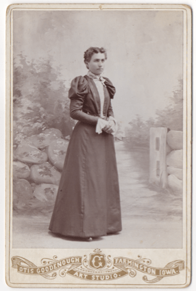 A woman wearing a full dress with lace cuffs and collar. Her hands are folded at her waist. Her hair dark and pulled back. It is curly in the front and appears to be parted in the middle.