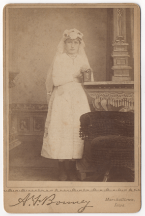A young girl dress in a white first confession dress. These outfits have a similar appearance to wedding dresses. She is clutching a small prayer book or Bible.