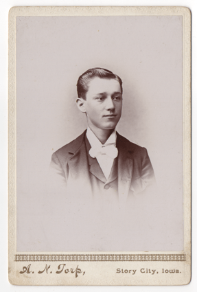 A dapper young man in formal wear. He has a dark suit and a white shirt with a large bow tie. His hair is short, parted on the left, and he has no facial hair.