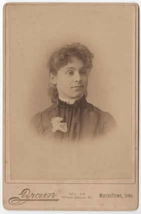 A curly haired woman with her hair swept back. She is wearing a pleated dress with a white lace flower on her lapel. She has a stiff white collar. 