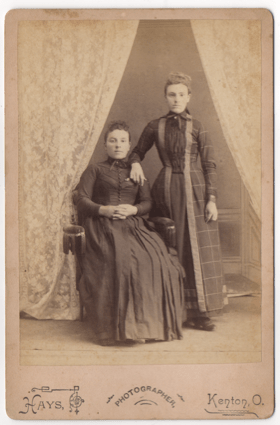 Two women. One seated, the other standing with her arm resting on the other's shoulder. Both wear full length dresses with one being a simpler dark dress with buttons down the front. The other with pleats and a pattern.