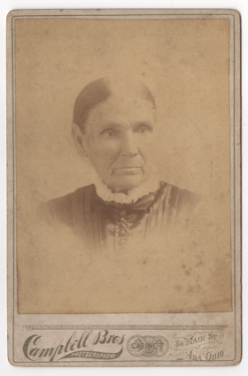 An old lady in a pleated dark dress with a frilly collar. Buttons adorn the front of the dress. Her hair is tightly pulled back and is parted down the center. The image is slightly faded, but it appeals she has pierced ears.
