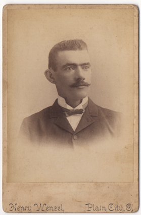A dapper gentleman in a bowtie and a dark suit. His undershirt is light colored and his color is upturned. His hair is well groomed and short in a buzzcut style. He has a mustache that is turned up at the ends.