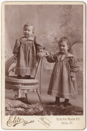 Two small children in matching dresses. One is standing in a chair, the other beside. The younger of the two appears to be a boy, the other a girl. Both dresses are embroidered and have decorative stitching.