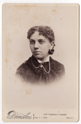 A young woman with dark curly hair parted down the middle. She is wearing a pendant style earring. She is wearing a dress with a light colored scalloped collar. She has a birch of clasp with a hanging stone and chains.