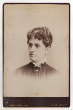 A woman with her hair piled on top of her head, pulled back, and pinned into place. She has a light colored ruffled collar, a dark pleated dress with buttons down the front, and a pendant earring. A clasp is at her throat.