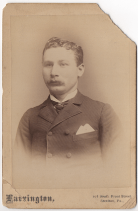 A dapper young man in a textured lined suit jacket and tie with some sort of pin through it. His hair is parted on the left and fairly short. He is looking slightly off camera. He has a mustache.