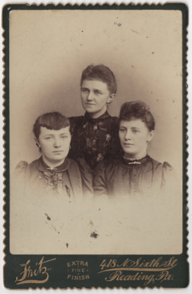 Three young women all in formal dresses. Two have hair curled in the front, one has bangs, all have their hair pulled back. All have their ears pierced and wear pendant or loop earrings. Various other pieces of jewelry are in the photo.