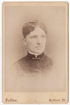 An older woman with her wavy hair pinned to her head and pulled back. She is wearing a dark dressed adorned with elaborate piping on either side of a centerline of large matching buttons. She has a white round collar. There is a clasp at her throat.