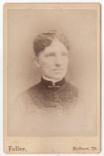 An older woman with her wavy hair pinned to her head and pulled back. She is wearing a dark dressed adorned with elaborate piping on either side of…